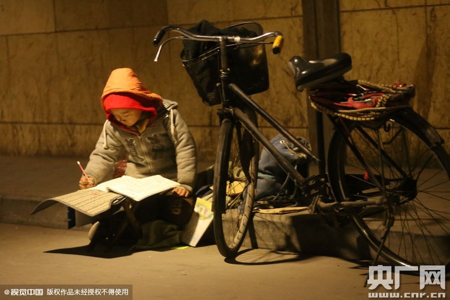 鄭州中原路京廣鐵路大橋下涵洞的馬路牙子邊，一位戴著紅色辣椒帽的小男孩趴在一個(gè)小馬扎上，上面鋪著一張車墊，在微弱的燈光下寫作業(yè)